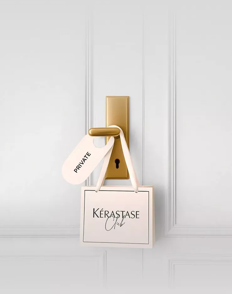 Une jeune femme aux cheveux bruns fonces, vetue d'un haut creme a decoupes et d'un pantalon clair, est allongee sur un fond gris clair, regardant directement l'objectif. Elle tient une carte de fidelite Kerastase dans sa main.