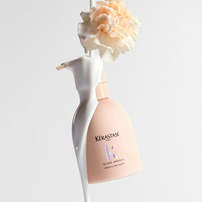 Flacon de creme Kerastase Gloss Absolu Frizz-Glaze avec un liquide blanc cremeux qui coule dessus et une fleur d'oeillet pale au sommet.