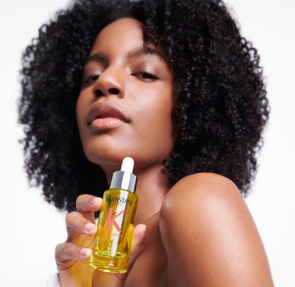Ebonee Davis, avec ses magnifiques cheveux bouclés, tient la meilleure huile pour cheveux abîmés Première Huile Gloss Réparatrice.