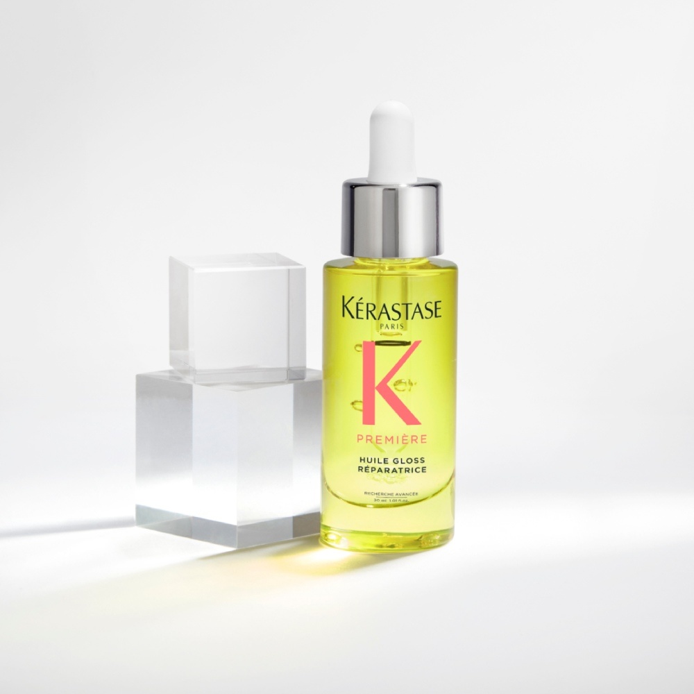 L'huile capillaire Première Huile gloss réparatrice est présentée sur un fond gris pur avec des cubes transparents. L'huile apporte de la brillance et scelle les pointes fourchues tout en neutralisant le ternissement.