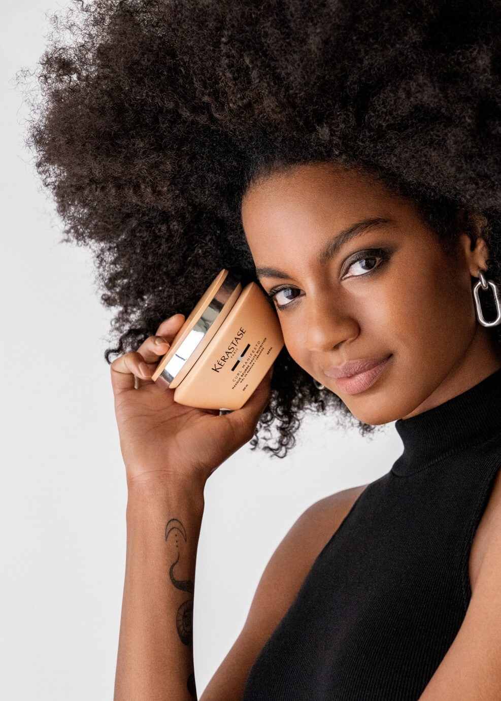 Femme souriante avec de cheveux bouclés tenant un pot de crème capillaire, vêtue d'un haut noir.