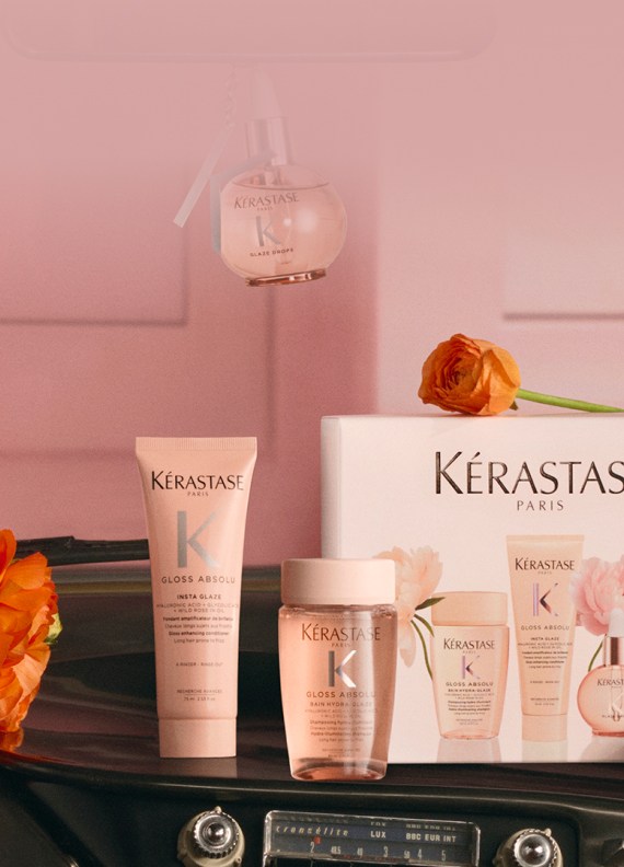 Image de produits capillaires Kerastase disposés pour un cadeau, incluant plusieurs flacons dorés, une bougie parfumée et une pochette blanche en tissu éponge, le tout présenté dans un emballage festif blanc et doré sur un fond blanc et lumineux. L'ensemble évoque le luxe et l'élégance.