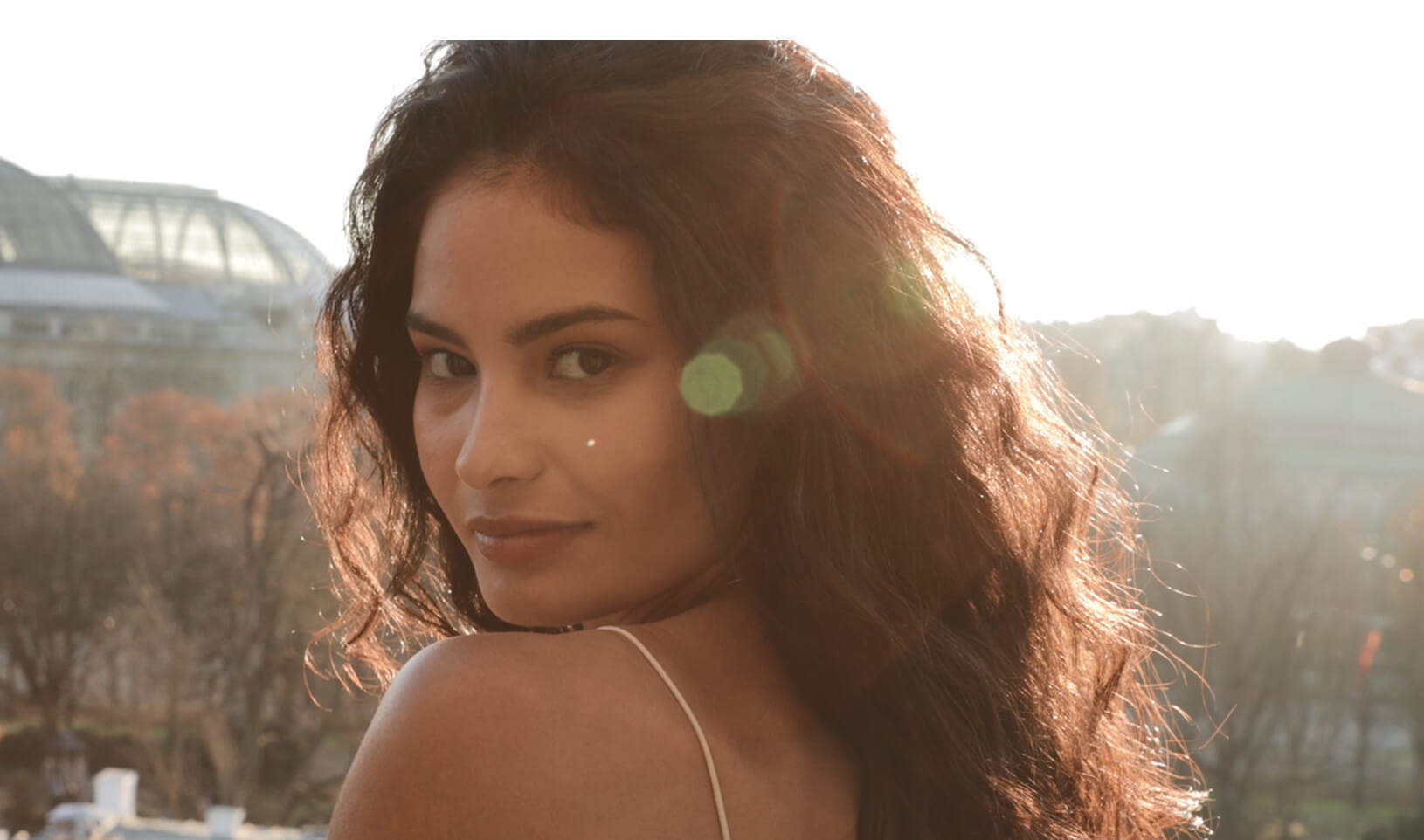 Une jeune femme au look naturel, cheveux bouclés, se tourne vers l'objectif avec un arrière-plan lumineux.