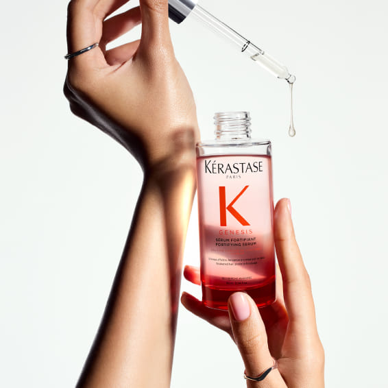Gros plan sur des mains tenant une bouteille de Kérastase Genesis Sérum Fortifiant, avec une pipette d'où s'apprête à tomber une goutte de produit.
