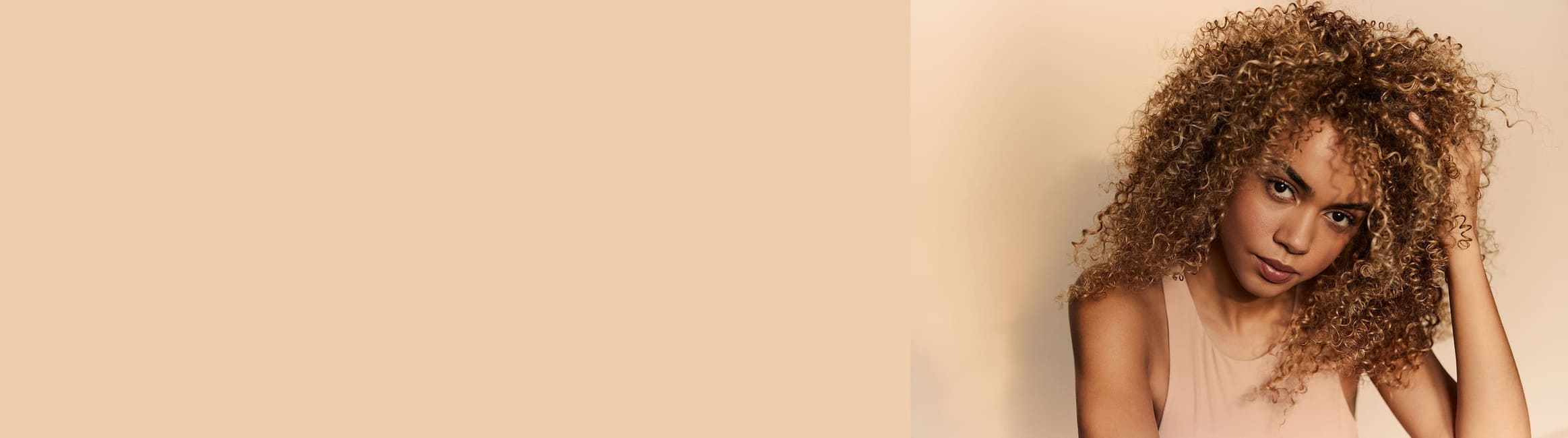 Une femme aux cheveux blonds bouclés et volumineux pose devant un fond de couleur crème. Elle regarde la caméra avec une expression pensive, la main légèrement posée sur ses cheveux.