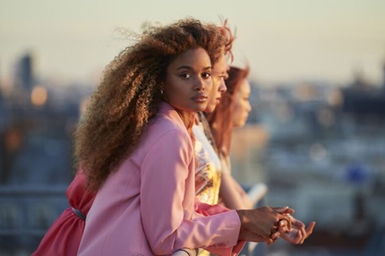 Trois femmes posent sur un toit, avec une vue urbaine en arrière-plan, illuminées par la lumière dorée du coucher de soleil.