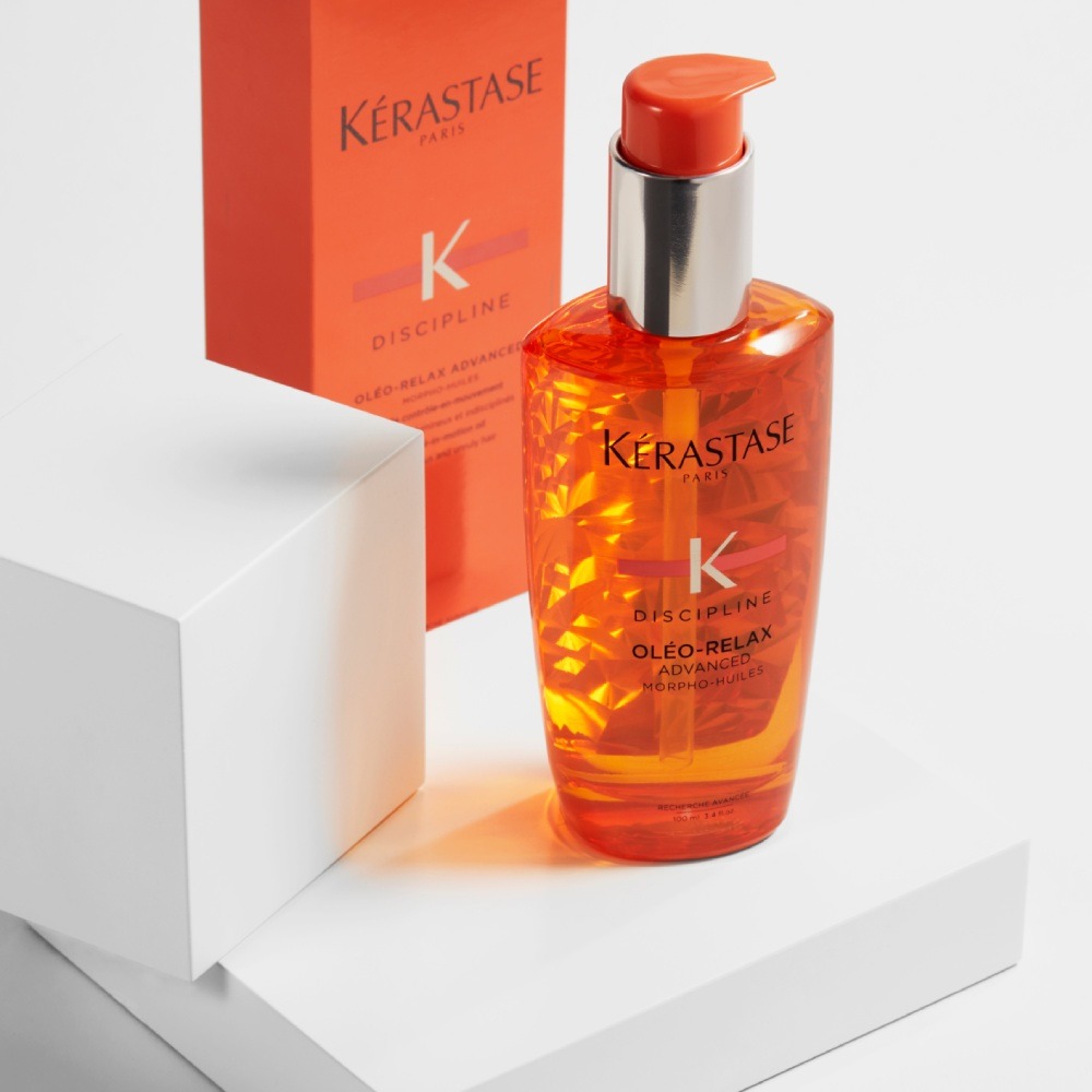 L'huile capillaire Discipline Oléo-Relax est présentée sur un fond gris pur avec des cubes. L'huile capillaire contrôle les frisottis, hydrate les cheveux et dompte leur volume.