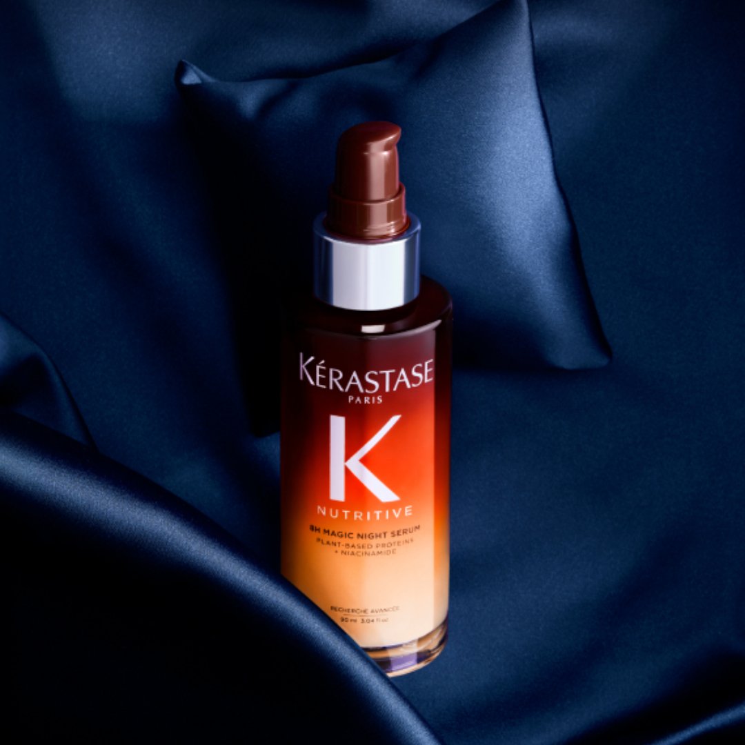 Trois flacons de produits capillaires Kérastase (Elixir Ultime, Genesis et Nutritive) sont disposés sur une surface blanche à côté d'un appareil photo vintage et de plusieurs pivoines blanches aux feuilles vertes. Le flacon doré Elixir Ultime est à gauche avec une pompe noire, le flacon rose Genesis avec un compte-gouttes argenté est au centre, et le flacon dégradé rouge-orangé Nutritive avec un compte-gouttes argenté est à droite. L'appareil photo argenté avec sa dragonne noire se trouve à gauche des produits.